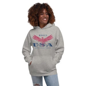 Może przedstawiać: Szara bluza z kapturem z nadrukiem z przodu. Grafika przedstawia stylizowanego orła z napisem "1913" powyżej i "USA NATIONAL TEAM" poniżej w kolorze czerwonym i niebieskim. Bluza ma kaptur i przednią kieszeń.