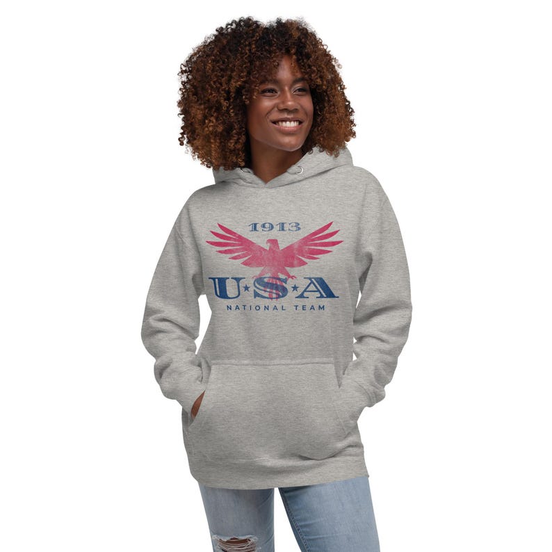 Może przedstawiać: Szara bluza z kapturem z nadrukiem z przodu. Grafika przedstawia stylizowanego orła z napisem "1913" powyżej i "USA NATIONAL TEAM" poniżej w kolorze czerwonym i niebieskim. Bluza ma kaptur i przednią kieszeń.