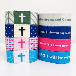 May include: A stack of four elastic fabric wristbands featuring cross symbols on the front and printed scripture text on the back. The bands include green, navy blue, pink, and light blue patterns with white, black, pink, and blue cross icons.