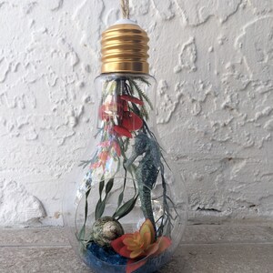 May include: A clear glass light bulb ornament with a gold metal base, filled with dried flowers, greenery, and a seashell. The ornament is hanging from a twine string.
