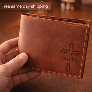 May include: Brown leather wallet with a cross design and the name "DAVID" engraved on the front. The wallet is held in a hand and appears to be new. The text "The Perfect Personalized Gift for Him" is at the top.