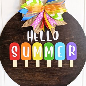 May include: A round wooden door hanger with the text "Hello Summer" in white letters. The letters are surrounded by six colorful popsicle shapes. A large bow made of colorful ribbons is attached to the top of the door hanger.