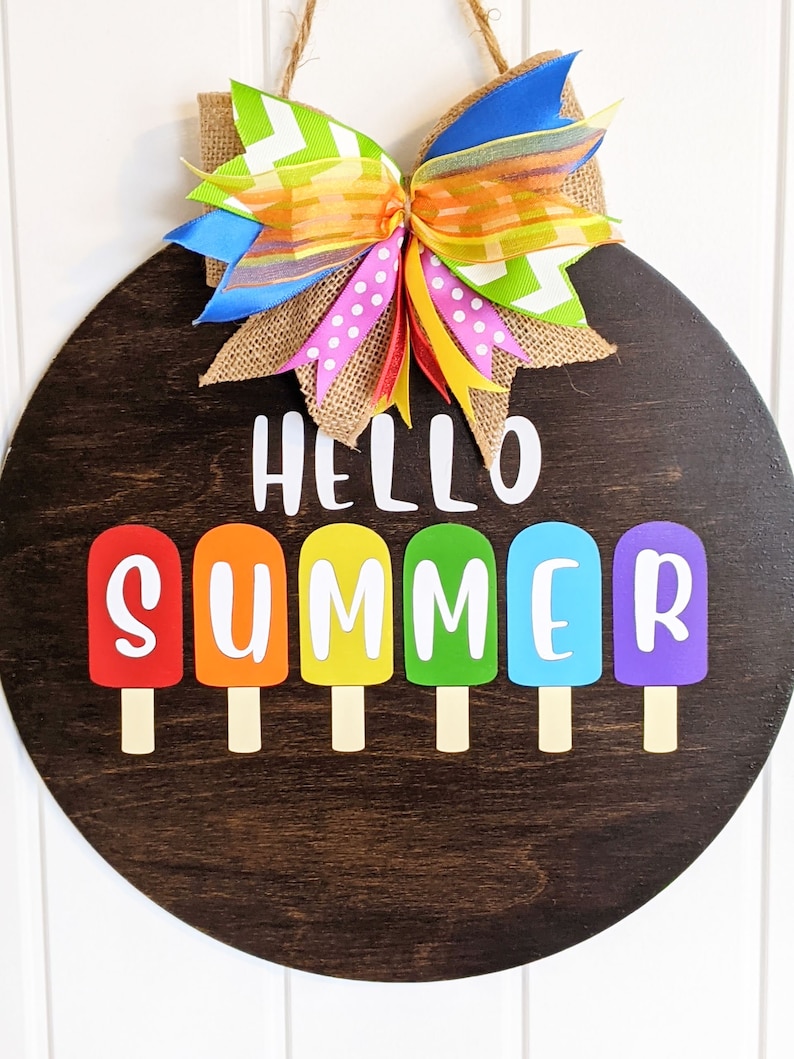 May include: A round wooden door hanger with the text "Hello Summer" in white letters. The letters are surrounded by six colorful popsicle shapes. A large bow made of colorful ribbons is attached to the top of the door hanger.
