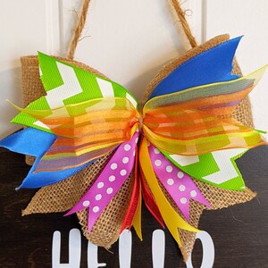 May include: A burlap wreath with a large bow made of colorful ribbons. The bow is tied with a red ribbon and has a yellow, orange, green, blue, and pink ribbon. The wreath has the words "HELLO" written on it in white.