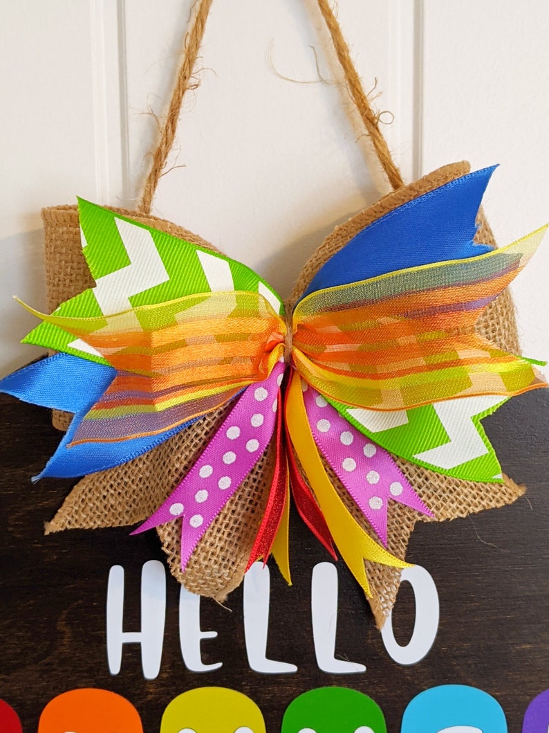 May include: A burlap wreath with a large bow made of colorful ribbons. The bow is tied with a red ribbon and has a yellow, orange, green, blue, and pink ribbon. The wreath has the words "HELLO" written on it in white.