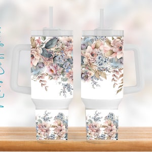 May include: Two white tumblers with a floral design. The design features pink, blue, and brown watercolor flowers. The tumblers have a white lid and a handle.