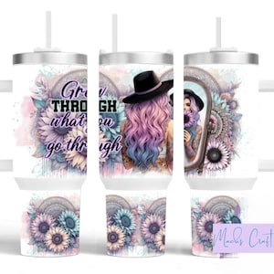 May include: White stainless steel tumbler with a handle and a straw. The tumbler has a design of a woman with pink and blue hair wearing a black hat and holding a sunflower. The text "Grow through what you go through" is on the tumbler.
