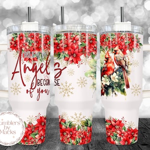 May include: White tumbler with a red and green floral design and two red cardinals. The tumbler has a handle and a straw. The text "I believe in angels because of you" is printed on the tumbler.