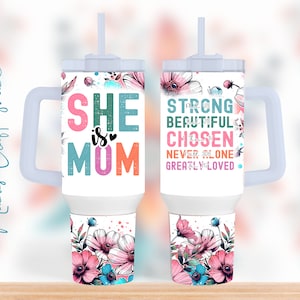 May include: Two white stainless steel tumblers with a clear lid and handle. The tumbler on the left has the text "SHE is MOM" in colorful letters with a pink heart. The tumbler on the right has the text "STRONG BEAUTIFUL CHOSEN NEVER ALONE GREATLY LOVED" in colorful letters. Both tumblers have a floral design with pink, blue, and green flowers.