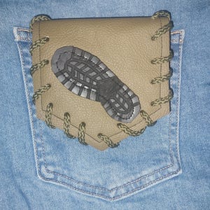 May include: A tan leather pocket cover featuring a dark gray boot print graphic in the center. The cover is secured with olive green cord lacing along the edges and is positioned over the back pocket of a pair of light blue denim jeans.