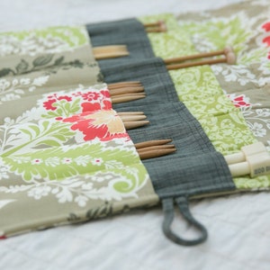 May include: A green, red, and white floral fabric roll-up case with a gray fabric flap. The case is filled with wooden knitting needles.