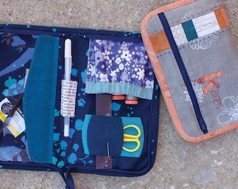 May include: Two fabric zippered pouches, one blue with white paint splatter and a clear pocket, the other gray with white floral embroidery and the name "Karen" embroidered in red. The blue pouch is open and contains sewing supplies, including a white pen, a ruler, scissors, and thread.