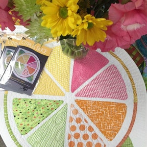 May include: A white quilted table runner with a citrus slice design. The runner is made of different fabrics with various patterns, including polka dots, stripes, and geometric designs. The runner is surrounded by a green border and has a white background.