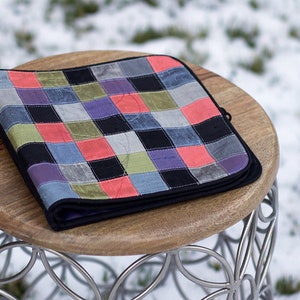 May include: A patchwork quilt with squares of various colors, including black, gray, blue, green, and pink, is folded and placed on a wooden stool with a silver metal base.