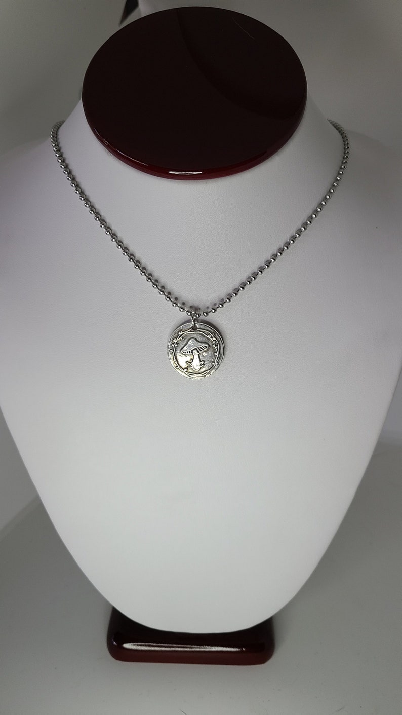 May include: A silver necklace with a round pendant featuring a detailed mushroom design. The pendant is surrounded by a delicate vine pattern.