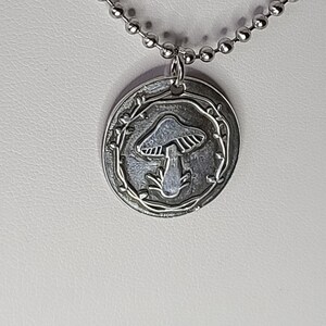 May include: A silver necklace with a round pendant featuring a mushroom design surrounded by a vine. The pendant is attached to a silver chain.