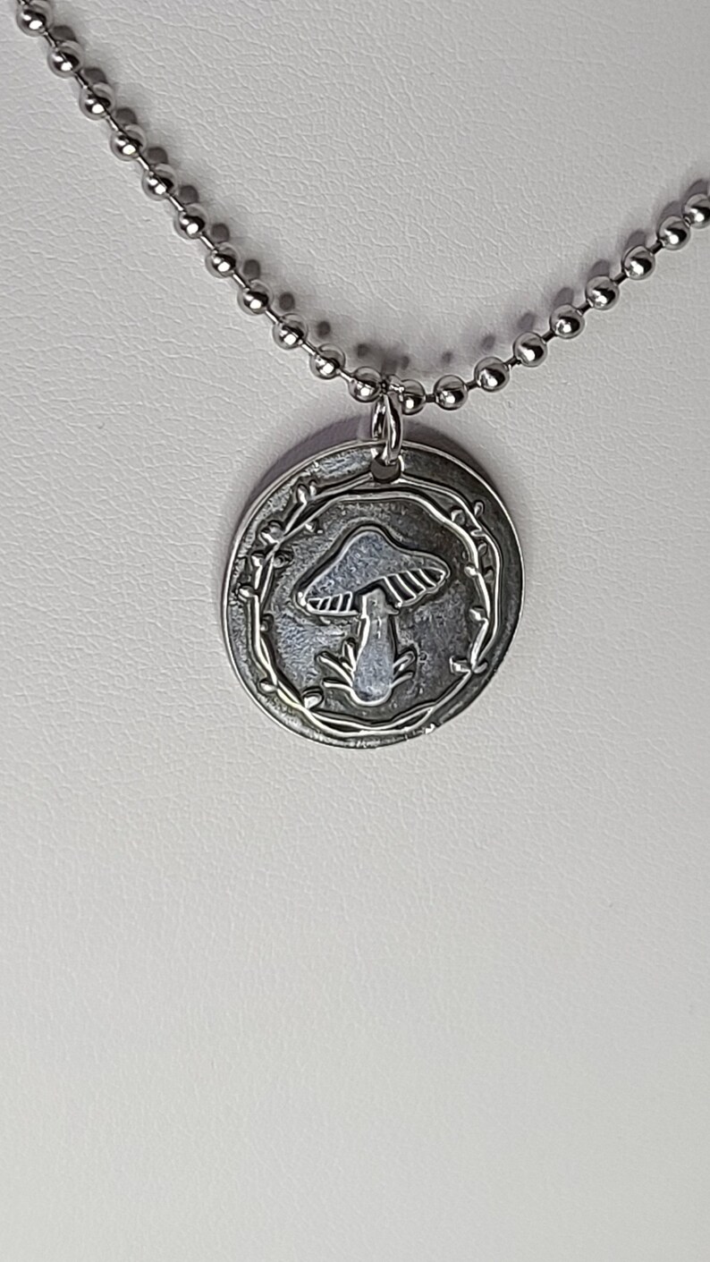 May include: A silver necklace with a round pendant featuring a mushroom design surrounded by a vine. The pendant is attached to a silver chain.