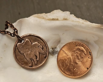 May include: A copper pendant necklace featuring a detailed silhouette of a dog, with a penny for scale. The pendant is on a chain and is displayed on a white seashell.