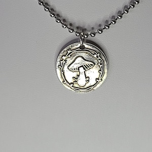 May include: A silver necklace with a round pendant featuring a mushroom design surrounded by a leafy wreath.