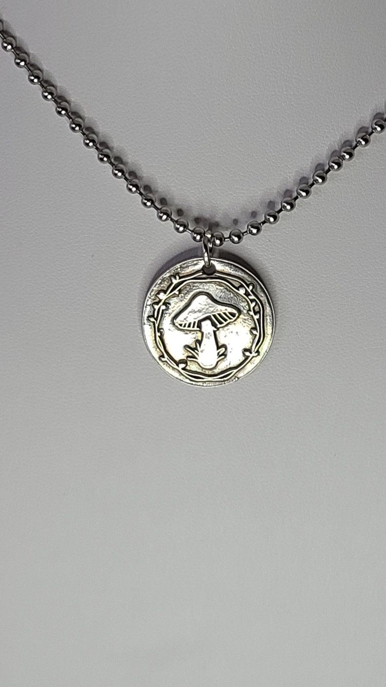 May include: A silver necklace with a round pendant featuring a mushroom design surrounded by a leafy wreath.