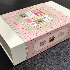 May include: A white rectangular gift box with a pink lid and a decorative band. The lid features a patchwork design with baby-themed illustrations and the words "Welcome Baby." The band has a heart pattern and the text "You Have My Heart."