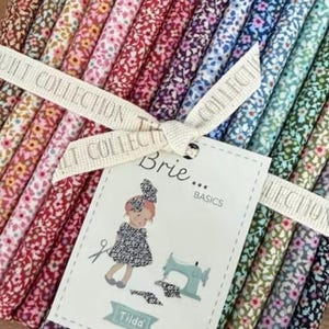 Peut inclure: Un assortiment de bandes de tissu aux motifs floraux variés dans un spectre de couleurs, noué par un ruban crème imprimé du texte QUILT COLLECTION. Une étiquette blanche affiche le texte Brie... BASICS, Tilda et une illustration.