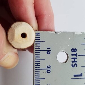 May include: A close-up of a wooden dowel with a hole drilled through the center. The dowel is being measured with a ruler, showing it is approximately 1 inch in length.