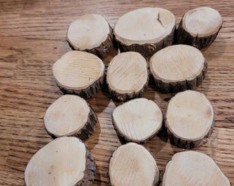 May include: A collection of small, round wooden slices with natural bark edges. The light-colored wood grain is visible on the flat surfaces. These rustic wood slices are arranged on a wooden surface.
