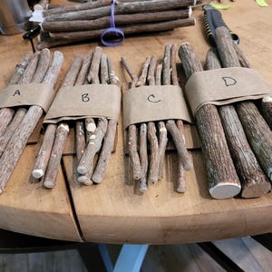 May include: Bundles of wooden sticks, labeled A, B, C, and D, are wrapped in brown paper. The sticks vary in thickness and have a natural, textured appearance. The ends of some sticks are cut flat. The image is taken on a wooden table.