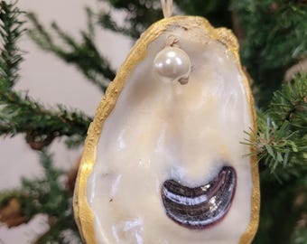 May include: A handcrafted oyster shell Christmas ornament. The shell is painted with gold trim and features a pearl and a dark, iridescent accent. A natural twine loop is attached for hanging. The ornament is displayed on a Christmas tree.