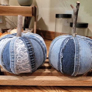 May include: Two decorative pumpkins crafted from strips of blue denim fabric. The pumpkin on the left features white lace panels between the denim sections. Both pumpkins have a natural wooden twig stem attached to the top center.
