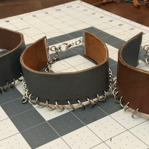 May include: Three leather chokers with silver chain accents. The chokers are in gray, brown, and black.
