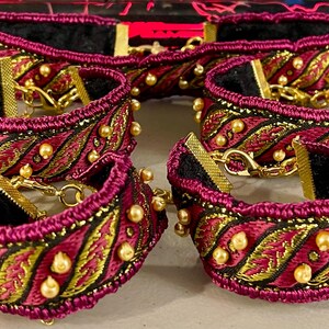 Könnte beinhalten: Fünf burgunderfarbene Stoffarmbänder mit goldenen Verzierungen und perlmuttartigen Akzenten. Die Armbänder sind durch eine goldene Kette verbunden.