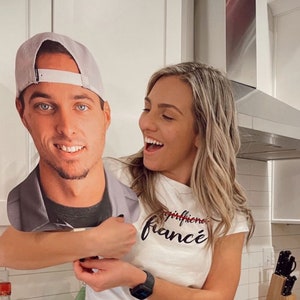 Puede incluir: Una mujer con una camiseta blanca con el texto "girlfriend fianc&eacute;" en letras negras sostiene un recorte de cart&oacute;n de un hombre con una gorra de b&eacute;isbol gris. El hombre sonr&iacute;e y mira a la c&aacute;mara. La mujer se r&iacute;e y mira al hombre. El fondo es una cocina con gabinetes blancos y una estufa negra.