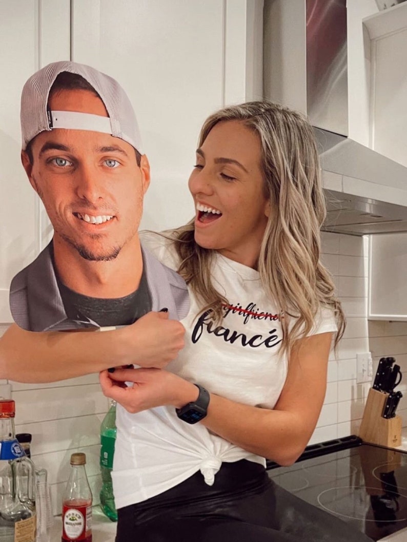 Puede incluir: Una mujer con una camiseta blanca con el texto "girlfriend fianc&eacute;" en letras negras sostiene un recorte de cart&oacute;n de un hombre con una gorra de b&eacute;isbol gris. El hombre sonr&iacute;e y mira a la c&aacute;mara. La mujer se r&iacute;e y mira al hombre. El fondo es una cocina con gabinetes blancos y una estufa negra.