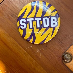 May include: A circular pin-back button featuring a yellow and purple tiger stripe pattern. The letters STTDB are printed in a bold, white, block-style font across the center of the button, overlaying the striped background design.