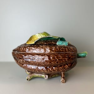 Puede incluir: Una fuente de cerámica marrón con forma de vaina de cacao, con una superficie texturizada. La tapa está adornada con detalles de hojas verdes y doradas. La fuente descansa sobre tres pequeñas patas con detalles verdes.