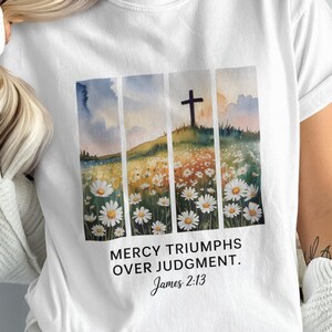May include: White t-shirt featuring a watercolor design of a cross on a hill, daisies, and a field. The text "MERCY TRIUMPHS OVER JUDGMENT. James 2:13" is printed below the image. The shirt is a casual, everyday wear item.
