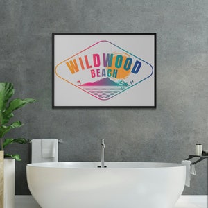 May include: A framed print featuring a colourful graphic design of a diamond shape with the text "WILDWOOD BEACH" inside. The graphic depicts a sunset, palm trees, and a wave. The print is hanging on a grey concrete wall above a white bathtub.