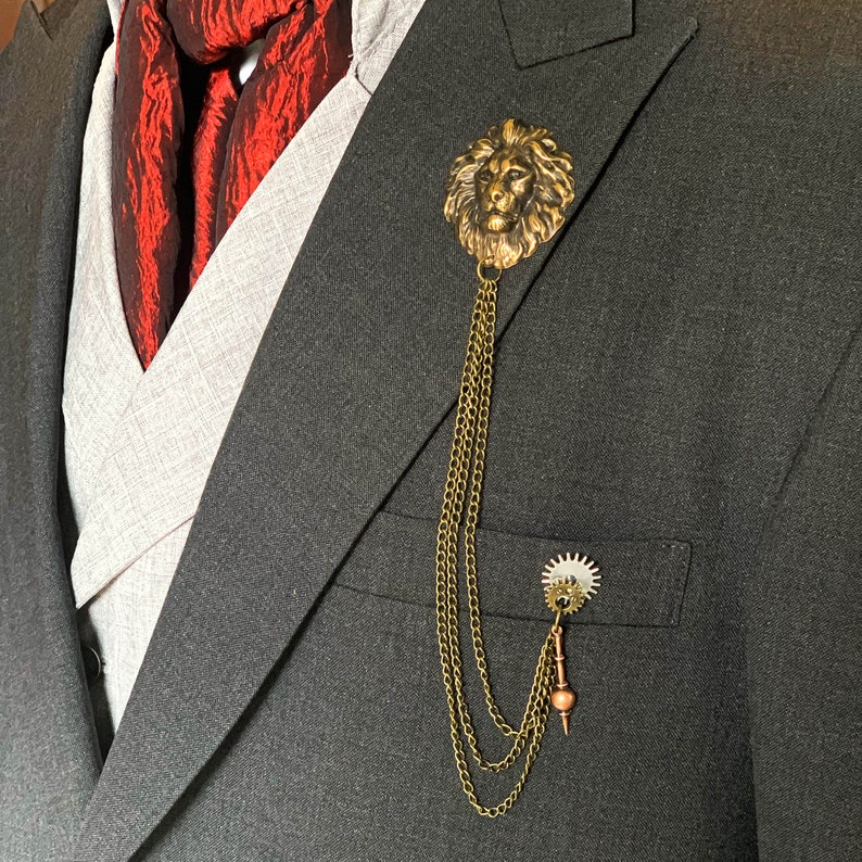 K&ouml;nnte beinhalten: Eine goldene Anstecknadel mit einem L&ouml;wenkopf, einer Kette und einem Zahnrad-Anh&auml;nger. Die Nadel ist an einem dunkelgrauen Anzugjackett befestigt.
