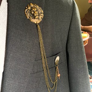 K&ouml;nnte beinhalten: Eine goldene Anstecknadel mit einem L&ouml;wenkopf, an der eine Kette befestigt ist, die mit einer kleineren, zahnradartigen Nadel verbunden ist. Die Nadel wird an einem dunkelgrauen Anzugjackett getragen.