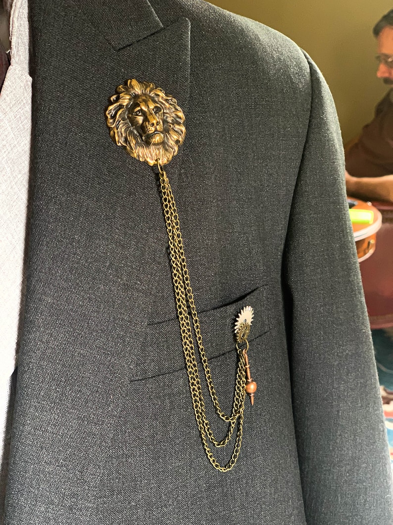 K&ouml;nnte beinhalten: Eine goldene Anstecknadel mit einem L&ouml;wenkopf, an der eine Kette befestigt ist, die mit einer kleineren, zahnradartigen Nadel verbunden ist. Die Nadel wird an einem dunkelgrauen Anzugjackett getragen.
