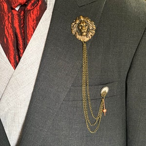 K&ouml;nnte beinhalten: Eine goldene Anstecknadel mit einem L&ouml;wenkopf und einer daran befestigten Kette. Die Nadel wird an einem grauen Anzugjackett getragen.