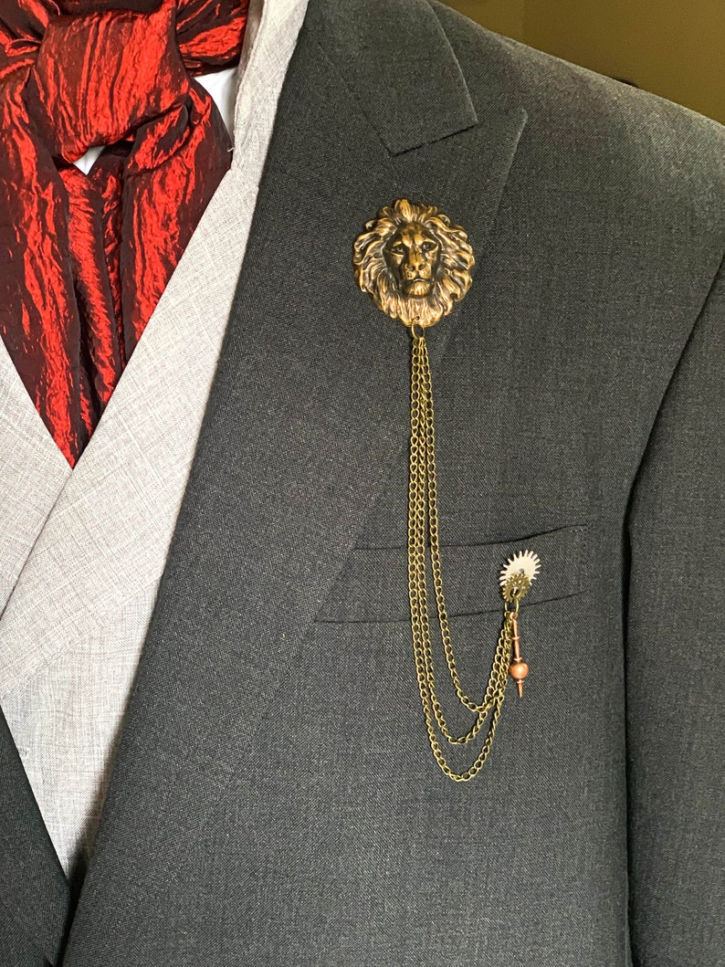 K&ouml;nnte beinhalten: Eine goldene Anstecknadel mit einem L&ouml;wenkopf und einer daran befestigten Kette. Die Nadel wird an einem grauen Anzugjackett getragen.