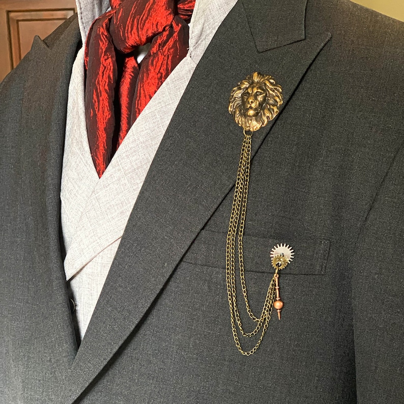 K&ouml;nnte beinhalten: Eine goldene Anstecknadel mit einem L&ouml;wenkopf und einer daran befestigten Kette. Die Nadel wird an einem dunkelgrauen Anzugjackett mit einem roten gemusterten Schal getragen.