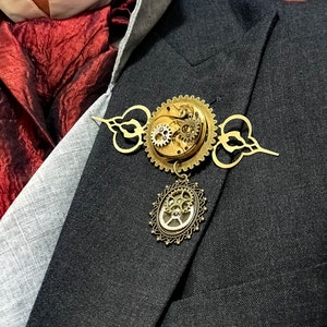 May include: A gold-toned steampunk-style brooch with intricate gears and a dangling circular charm. The brooch is pinned to a gray suit jacket.