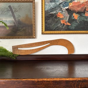 May include: A vintage wooden dressmaker's curve ruler with a curved design and measurement markings. The ruler is displayed on a dark wooden surface, with framed artwork in the background.