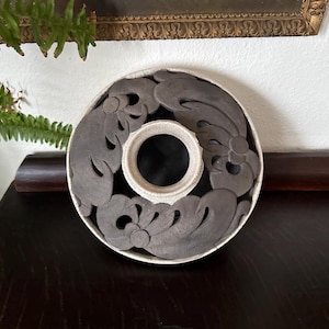 May include: A circular, decorative ceramic piece with a central hole. The piece features a dark gray, floral-like design on a white and gray background. The object is placed on a dark wooden surface.