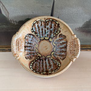May include: A round ceramic bowl with two side handles, featuring a beige base color and a central floral-like pattern in shades of brown, blue, and yellow. The interior surface has a textured, glazed finish with distinct, raised petal-shaped sections.