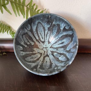 May include: A round ceramic bowl featuring a dark charcoal base color with a reactive glaze pattern in shades of light blue and white that radiates from the center in a petal-like design. The interior surface has a glossy, smooth finish.